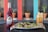 A vibrant bottle of strawberry Jarritos soda is placed next to a plate with three tacos topped with shredded purple cabbage and avocado sauce. Beside the plate is a glass jar with what appears to be seasoned fries or sticks. In the background, there are blurred potted plants against a colorful backdrop.