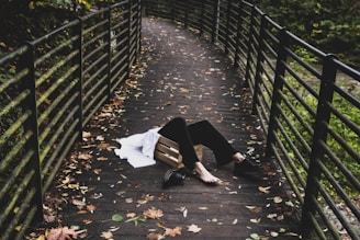 A person lies on a wooden pedestrian bridge surrounded by metal railings, with autumn leaves scattered across the ground. The person appears to be in a relaxed or vulnerable position, wearing black pants and black shoes, with a white garment nearby.