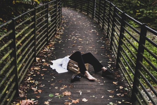 A person lies on a wooden pedestrian bridge surrounded by metal railings, with autumn leaves scattered across the ground. The person appears to be in a relaxed or vulnerable position, wearing black pants and black shoes, with a white garment nearby.