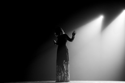 A person stands on stage under dramatic lighting, casting a shadow against a dark background. The light creates a striking silhouette, highlighting the figure's outstretched hand and long dress.