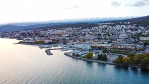An aerial view of a coastal city with numerous buildings, a marina filled with boats, and lush greenery along the shoreline. The city is nestled against a backdrop of rolling hills and a picturesque body of water in the foreground.