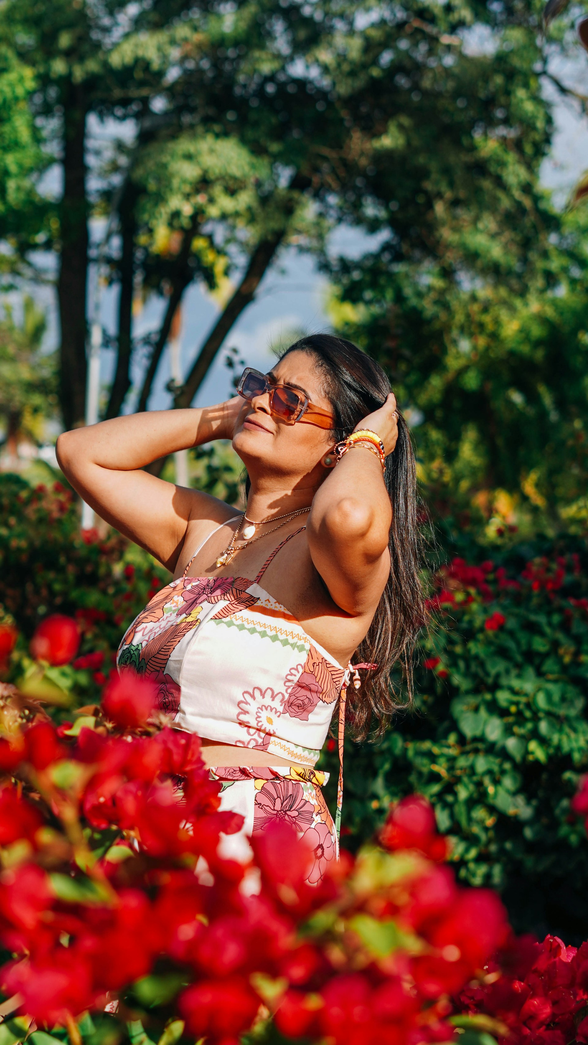 A stylish woman wearing a flowing summer dress stands in a sunlit park, surrounded by blooming flowers.