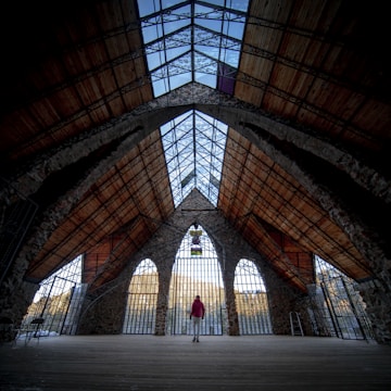 A large, open architectural interior with high, vaulted stone arches and a wooden ceiling. The structure features expansive glass windows, allowing in natural light. A person wearing a red jacket stands in the center, adding a sense of scale. The design conveys a blend of rustic and modern elements.