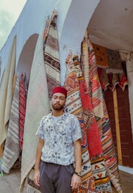 A man wearing a patterned shirt and a red hat stands in front of vibrant, intricately designed rugs hanging from an archway. The rugs display various colorful geometric and tribal patterns.