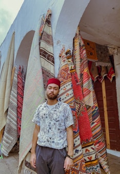 A man wearing a patterned shirt and a red hat stands in front of vibrant, intricately designed rugs hanging from an archway. The rugs display various colorful geometric and tribal patterns.