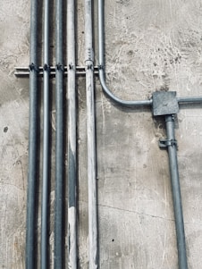 Multiple metal pipes are mounted along a textured concrete wall. The pipes vary in size and are arranged both vertically and horizontally, connected by clamps and a junction box.