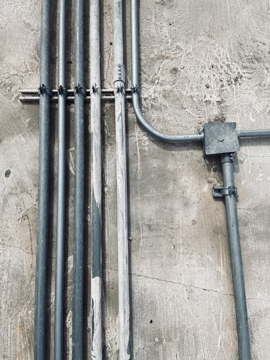 Multiple metal pipes are mounted along a textured concrete wall. The pipes vary in size and are arranged both vertically and horizontally, connected by clamps and a junction box.