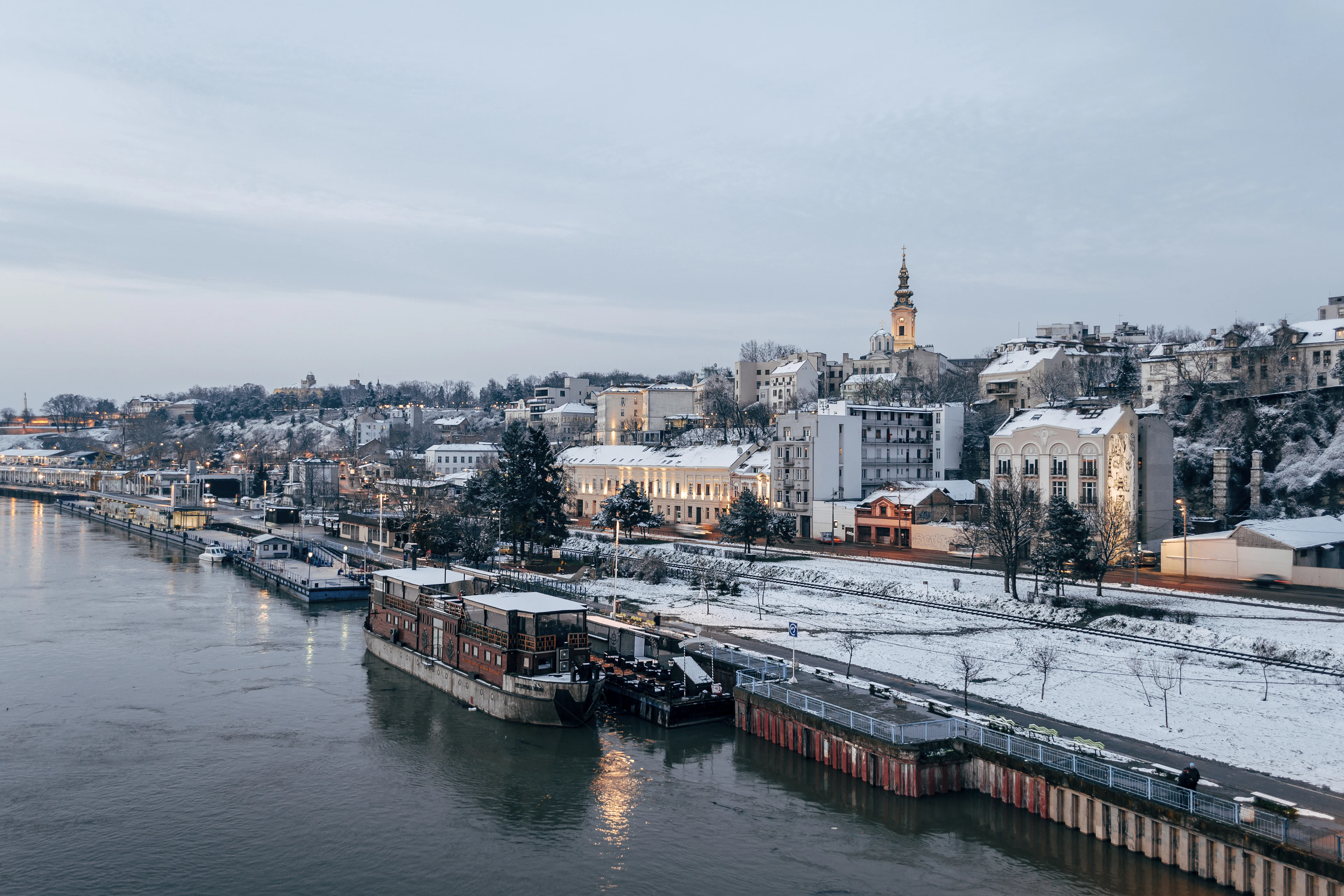 Snow along the Sava quay in Belgrade, floating restaurants moored beneath the frosted rooftops and a clock tower of the old town