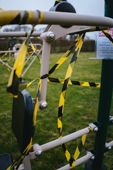 An outdoor gym machine wrapped in yellow and black caution tape suggesting it is off-limits. A sign indicates temporary closure due to COVID restrictions. The equipment is situated on grass with a blurred background of more gym equipment.