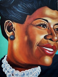 A vibrant mural featuring a detailed portrait of a woman with an elegant pose, highlighted by a distinctive smile and bold red lipstick. She wears a large pearl necklace and matching earrings, and her hair is styled in a classic updo. The background is a smooth teal color, which contrasts with her deep skin tone.