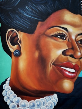 A vibrant mural featuring a detailed portrait of a woman with an elegant pose, highlighted by a distinctive smile and bold red lipstick. She wears a large pearl necklace and matching earrings, and her hair is styled in a classic updo. The background is a smooth teal color, which contrasts with her deep skin tone.