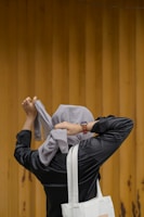 A person dressed in a black jacket is tying a light gray headscarf against a wooden planked background. A white tote bag hangs from their shoulder, and they are wearing a watch on their left wrist.