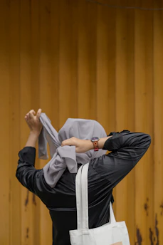 A person dressed in a black jacket is tying a light gray headscarf against a wooden planked background. A white tote bag hangs from their shoulder, and they are wearing a watch on their left wrist.