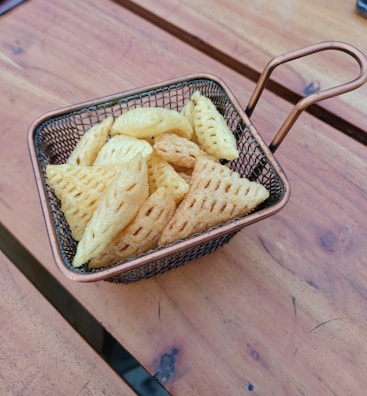 A small wire basket filled with triangular crispy snacks is placed on a wooden table. The snacks have a waffle-like texture and are golden brown in color.