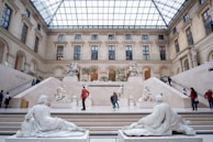 Visitors admiring priceless artworks inside a grand museum hall with high ceilings and ornate details.