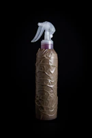 A spray bottle wrapped in crumpled brown paper is placed against a dark background. The bottle features a clear plastic trigger sprayer at the top.