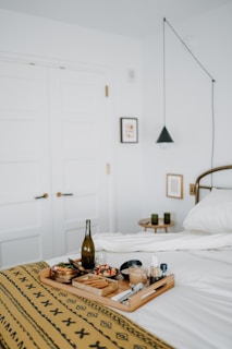 A neatly arranged breakfast tray rests on a bed covered with a patterned yellow blanket. The tray includes a bottle of what appears to be wine or juice, a latte, chopped fruit, a small dish, cutlery, and condiments. In the background, there are white double doors, a small framed picture on the wall, a black pendant light, and a side table with two green candles.
