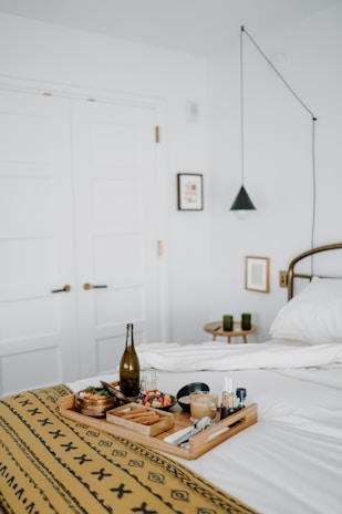 A neatly arranged breakfast tray rests on a bed covered with a patterned yellow blanket. The tray includes a bottle of what appears to be wine or juice, a latte, chopped fruit, a small dish, cutlery, and condiments. In the background, there are white double doors, a small framed picture on the wall, a black pendant light, and a side table with two green candles.