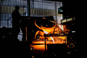 A close-up of hands shaping molten metal in an industrial foundry, glowing orange against dark machinery.
