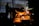 A silhouette of a worker stands near an industrial furnace in a dimly lit metalworks setting. Bright orange molten metal pours from a container, creating sparks. The background includes metal structures and equipment, indicating an industrial environment.