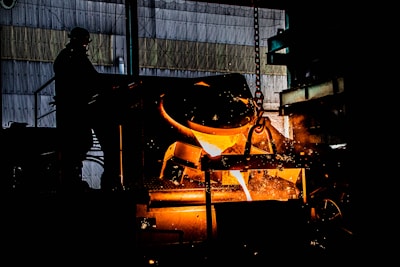 A silhouette of a worker stands near an industrial furnace in a dimly lit metalworks setting. Bright orange molten metal pours from a container, creating sparks. The background includes metal structures and equipment, indicating an industrial environment.