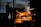 A silhouette of a worker stands near an industrial furnace in a dimly lit metalworks setting. Bright orange molten metal pours from a container, creating sparks. The background includes metal structures and equipment, indicating an industrial environment.