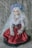 A doll with long white hair adorned with a floral crown sits against a backdrop of newspaper-like text. The doll is dressed in an elaborate outfit with a red skirt featuring black polka dots, a white frilled blouse, and decorative accessories, including pearl embellishments.