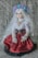 A doll with long white hair adorned with a floral crown sits against a backdrop of newspaper-like text. The doll is dressed in an elaborate outfit with a red skirt featuring black polka dots, a white frilled blouse, and decorative accessories, including pearl embellishments.