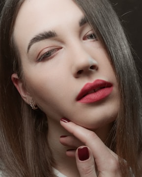 A close-up of a person's face with straight brown hair partially covering one eye and wearing red lipstick. The skin appears smooth and the eyebrows are neatly groomed. The person's hand with red painted nails is gently touching their chin, adding a contemplative or sensual element to the image. Small gold hoop earrings are worn on the ear. The background is blurred, drawing full attention to the face.
