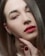 A close-up of a person's face with straight brown hair partially covering one eye and wearing red lipstick. The skin appears smooth and the eyebrows are neatly groomed. The person's hand with red painted nails is gently touching their chin, adding a contemplative or sensual element to the image. Small gold hoop earrings are worn on the ear. The background is blurred, drawing full attention to the face.