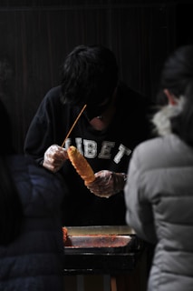A person wearing a black sweater with 'TIBET' written on it is preparing food on a skewer. They are using gloves and working behind a counter. Two other individuals are partially visible, positioned in the foreground.