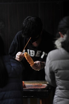 A person wearing a black sweater with 'TIBET' written on it is preparing food on a skewer. They are using gloves and working behind a counter. Two other individuals are partially visible, positioned in the foreground.