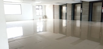 A spacious and well-lit empty room with a glossy tiled floor, featuring three elevator doors on one side. The walls are white and there are a couple of large windows letting in natural light.