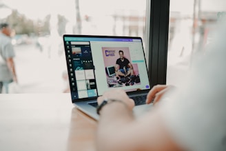 A person is using a laptop at a wooden table near a window. The screen displays an image editing software showing a photo of a smiling man with cameras and the text 'CONTENT IS CHAOS'. The view outside the window is slightly blurred, with a few people walking by.
