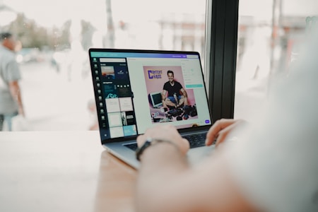 A person is using a laptop at a wooden table near a window. The screen displays an image editing software showing a photo of a smiling man with cameras and the text 'CONTENT IS CHAOS'. The view outside the window is slightly blurred, with a few people walking by.