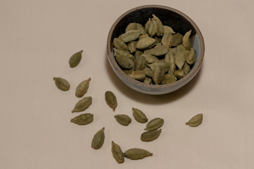 A ceramic bowl filled with green cardamom pods is placed on a beige surface. Several cardamom pods are scattered around the bowl on the surface, creating a simple, natural arrangement.
