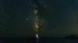 A night sky filled with stars and the Milky Way galaxy, casting a gentle glow over a solitary rock formation rising from the dark waters below. The vast expanse of the universe is emphasized by the shimmering celestial bodies.