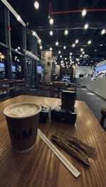 A cozy and modern coffee shop interior features warm lighting and a wooden table with a cup of coffee, sugar packets, and a straw. There are additional tables and chairs arranged neatly in the background, and a counter area with a visible menu.