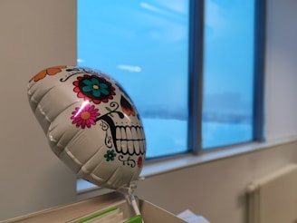A colorful, decorative balloon with a Day of the Dead sugar skull design floats in front of a large window. The skull is adorned with vibrant flowers and patterns, creating a festive look. Outside the window, an overcast sky suggests a winter setting.