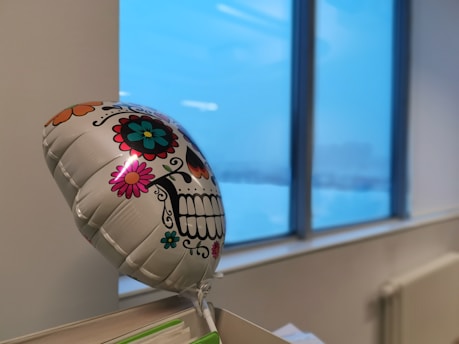 A colorful, decorative balloon with a Day of the Dead sugar skull design floats in front of a large window. The skull is adorned with vibrant flowers and patterns, creating a festive look. Outside the window, an overcast sky suggests a winter setting.