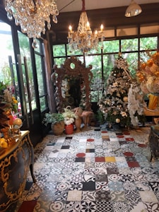 An elegantly decorated room is filled with ornate furnishings and decorations, including a large, detailed mirror with an intricate frame, a chandelier with hanging crystals, and a Christmas tree adorned with lights and ornaments. Various potted plants and flowers are arranged near the mirror, contributing to the room's lush and vibrant atmosphere. The floor is covered with decorative tiles featuring a mix of patterns and colors, adding a stylish and artistic touch to the space.