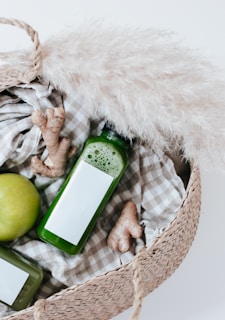 A woven basket contains a green apple, fresh ginger roots, and bottles of green juice. The basket is lined with a checkered fabric and is complemented by a piece of pampas grass.