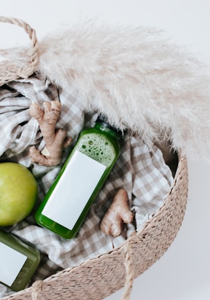 A woven basket contains a green apple, fresh ginger roots, and bottles of green juice. The basket is lined with a checkered fabric and is complemented by a piece of pampas grass.