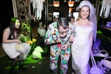 A group of people enjoying a festive occasion, possibly a wedding. The woman on the right is wearing a white wedding dress and veil, smiling joyfully. Next to her, a man in a colorful, patterned suit seems to be having fun. Another woman on the left is laughing and holding a basket filled with flower petals. The surroundings are elegantly decorated with floral arrangements and ornate decorations, contributing to a celebratory atmosphere.