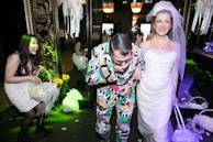 A group of people enjoying a festive occasion, possibly a wedding. The woman on the right is wearing a white wedding dress and veil, smiling joyfully. Next to her, a man in a colorful, patterned suit seems to be having fun. Another woman on the left is laughing and holding a basket filled with flower petals. The surroundings are elegantly decorated with floral arrangements and ornate decorations, contributing to a celebratory atmosphere.
