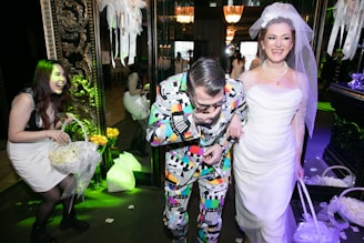 A group of people enjoying a festive occasion, possibly a wedding. The woman on the right is wearing a white wedding dress and veil, smiling joyfully. Next to her, a man in a colorful, patterned suit seems to be having fun. Another woman on the left is laughing and holding a basket filled with flower petals. The surroundings are elegantly decorated with floral arrangements and ornate decorations, contributing to a celebratory atmosphere.