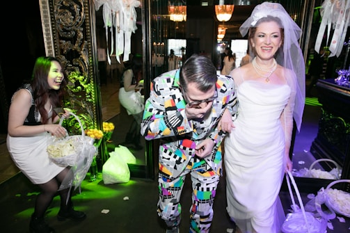 A group of people enjoying a festive occasion, possibly a wedding. The woman on the right is wearing a white wedding dress and veil, smiling joyfully. Next to her, a man in a colorful, patterned suit seems to be having fun. Another woman on the left is laughing and holding a basket filled with flower petals. The surroundings are elegantly decorated with floral arrangements and ornate decorations, contributing to a celebratory atmosphere.