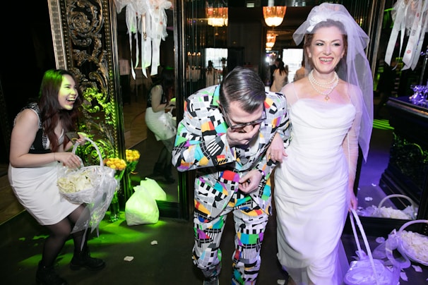 A group of people enjoying a festive occasion, possibly a wedding. The woman on the right is wearing a white wedding dress and veil, smiling joyfully. Next to her, a man in a colorful, patterned suit seems to be having fun. Another woman on the left is laughing and holding a basket filled with flower petals. The surroundings are elegantly decorated with floral arrangements and ornate decorations, contributing to a celebratory atmosphere.