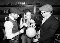 Three individuals are gathered around a globe, pointing and discussing its features. They are in a room with bookshelves, trophies, and other decor in the background. The attire suggests a vintage or formal theme, with flat caps and a vest.