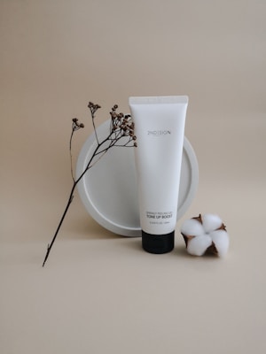 A minimalist still life featuring a white tube of skincare product labeled 'Springy Peeling Gel Tone Up Boost'. It is centered against a neutral beige background, accompanied by a sprig of small dried flowers and a piece of cotton. The arrangement conveys a sense of natural beauty and simplicity.